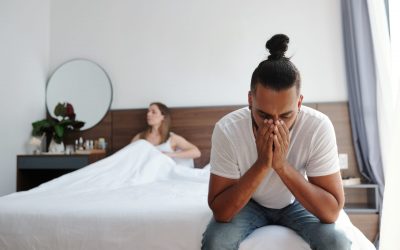 Stressed sad man sitting on bed suffering from erectile dysfunction, his upset wife is in background